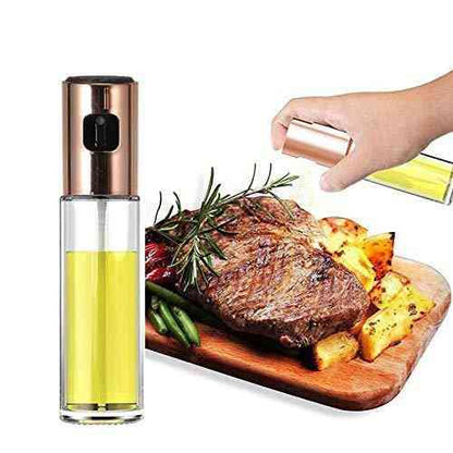 A clear glass oil dispenser spray bottle with a gold-colored lid, shown beside a wooden board with a steak and vegetables on it, indicating use for cooking.